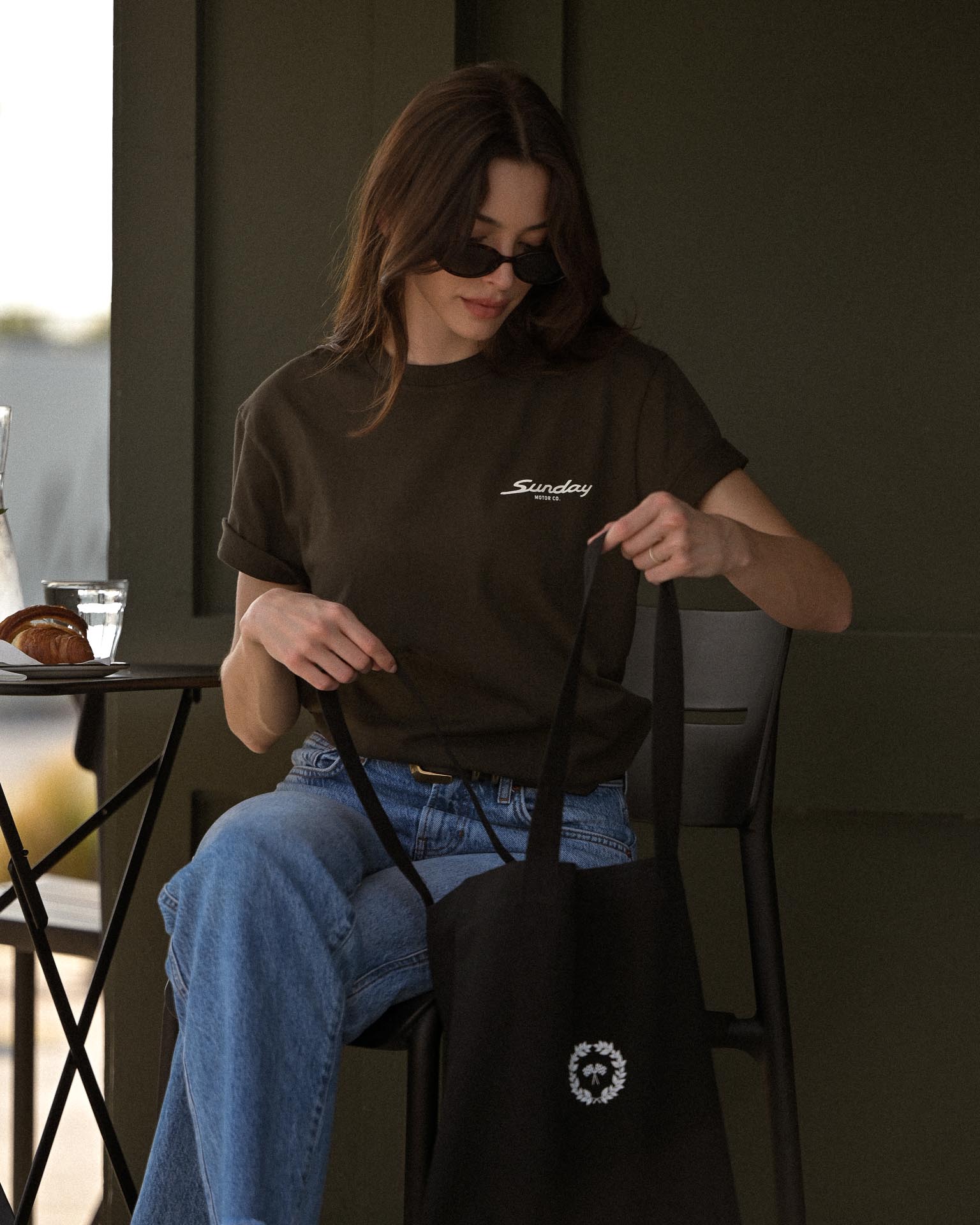 Woman sitting outdoors wearing a brown t-shirt with text and holding a black tote bag.